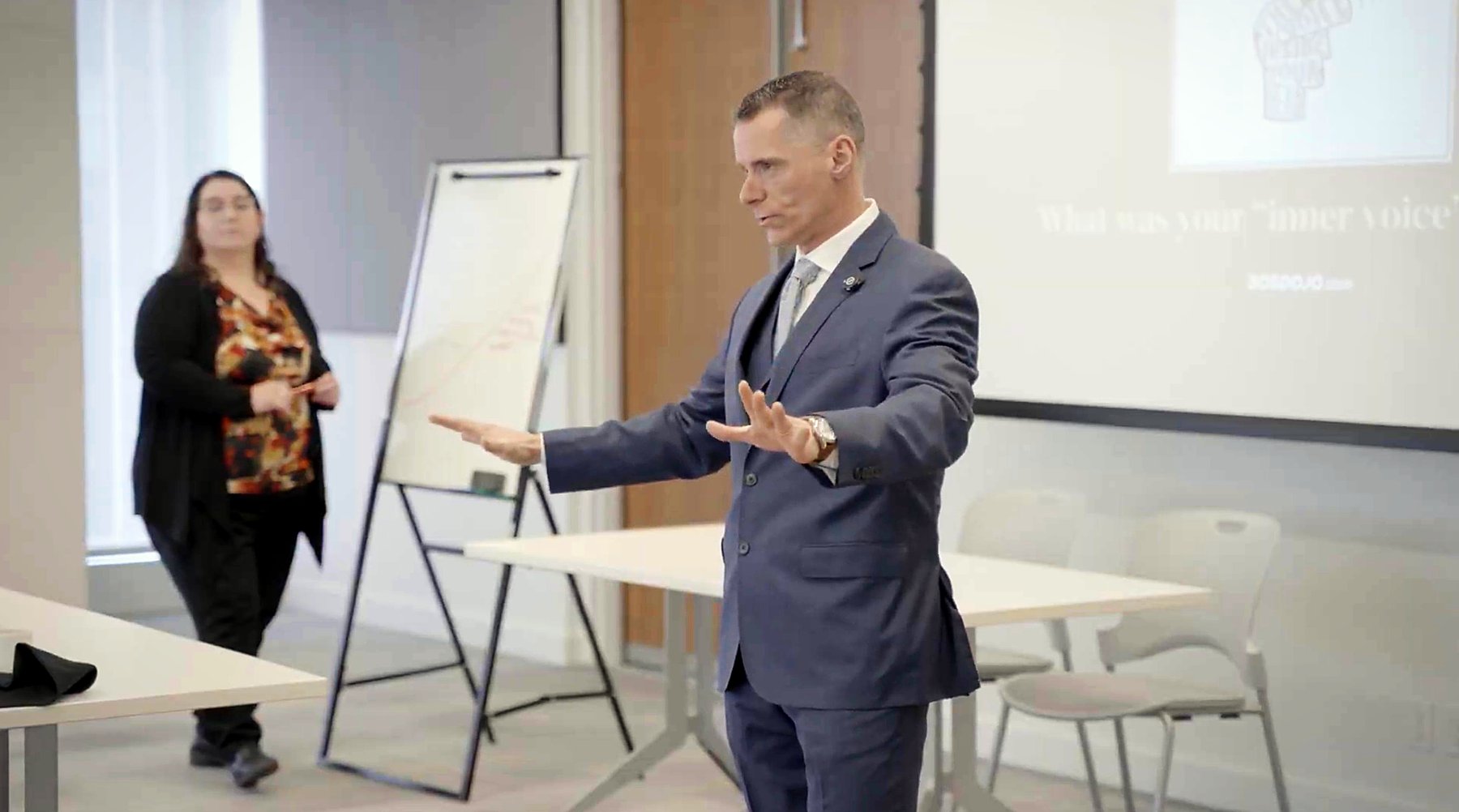 Joe Marcoux in a navy three-piece suit, mid-coaching gesture, leading an AON Insurance corporate training session. A participant stands at a flip chart in the background while a SOS Dojo slide reads 'What was your inner voice?' projected behind.