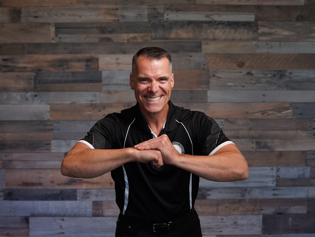 Joe Marcoux portrait, smiling, hands clasped, in a black SOS Dojo polo against a reclaimed-wood backdrop